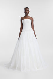 A model wearing a strapless white tulle wedding gown with a structured bodice and a voluminous, asymmetric layered tulle skirt, photographed against a seamless light gray studio background.