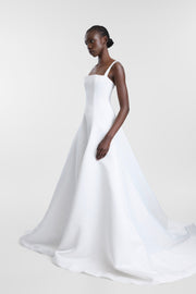 A woman modeling a white sleeveless A-line wedding gown with a fitted bodice and full skirt, photographed against a clean light gray studio background.