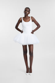 A model wearing a white sleeveless ballet leotard-style dress with a short, tutu skirt, paired with white pointed-toe heels, standing against a light gray studio background.