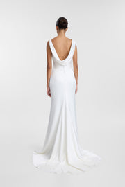 A woman viewed from the back wearing a white floor-length gown with a deep cowl neckline and a slight train, photographed against a light gray studio background.