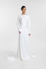A model wearing a long-sleeved white dress with a wide boat neckline, a fitted bodice, and a flowing skirt with a train, photographed against a seamless light gray studio background.