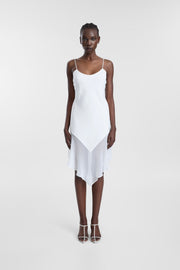 A model wearing a white spaghetti-strap slip dress with a draped, layered hemline, paired with white strappy high-heeled sandals, set against a plain light gray studio background.