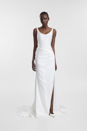 A model wearing a sleeveless white satin gown with a draped wrap-style skirt and a high front slit, standing against a light gray studio background.
