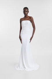 A woman modeling a strapless white mermaid-style gown with a fitted bodice, smooth draping, and a slight train against a plain light gray studio background.