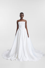 A model wearing a strapless white wedding corset with a structured bodice and a full, flowing skirt with a train, photographed against a light gray studio background.