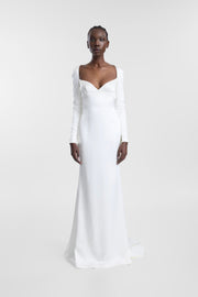 A model wearing a long-sleeved white wedding gown with a deep V-neckline and floor-length silhouette, photographed against a seamless light gray studio background.