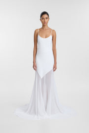 A model wearing a white spaghetti-strap slip dress with a structured bodice and a flowing, asymmetrical sheer skirt, photographed against a seamless light gray studio background.