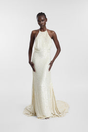 A model wearing a floor-length halter-neck gown covered in shimmering sequins, standing against a light gray studio background.
