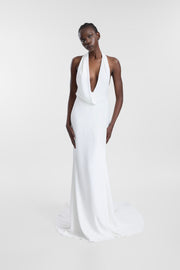 A model wearing a floor-length white halter gown with a plunging V-neckline and a slight train, photographed against a seamless light gray studio background.